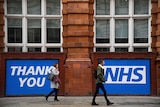 Two people walk in front of a large banner that reads "Thank you NHS"