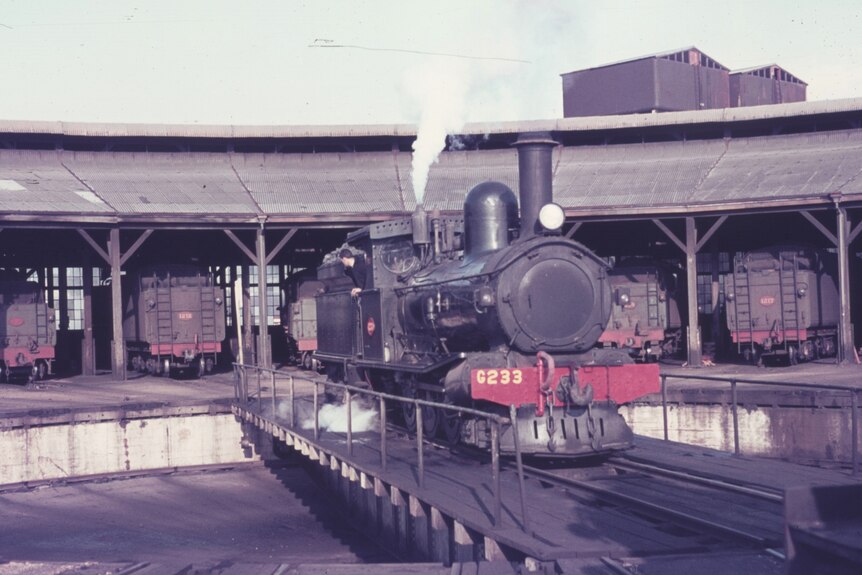 An old steam locomotive pictured outside a round house filled with train carriages.