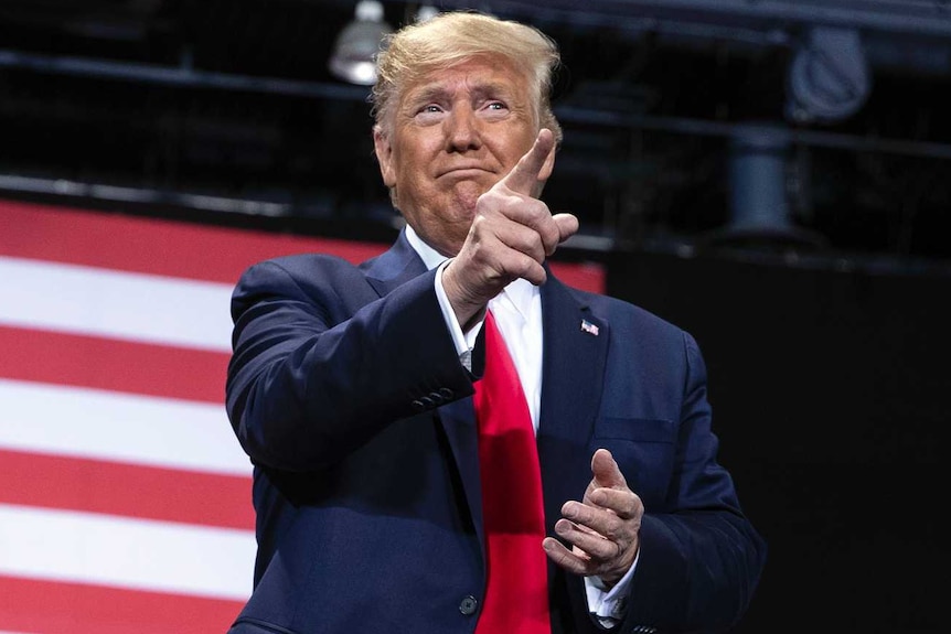 Donald Trump stands in front of a crowd and large flag at a rally, pointing