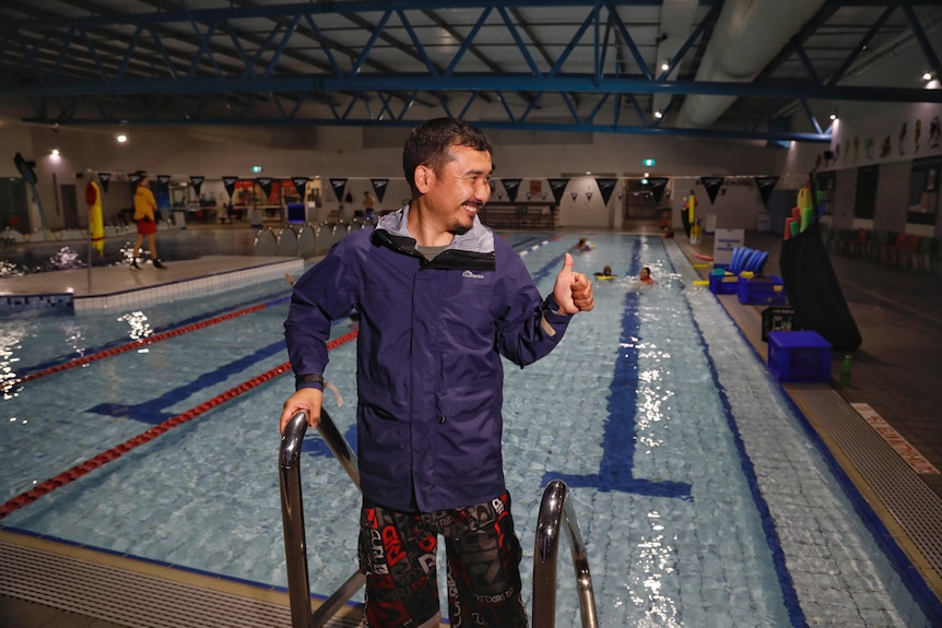 A man wearing a spray jacket and board shorts stands near the pool