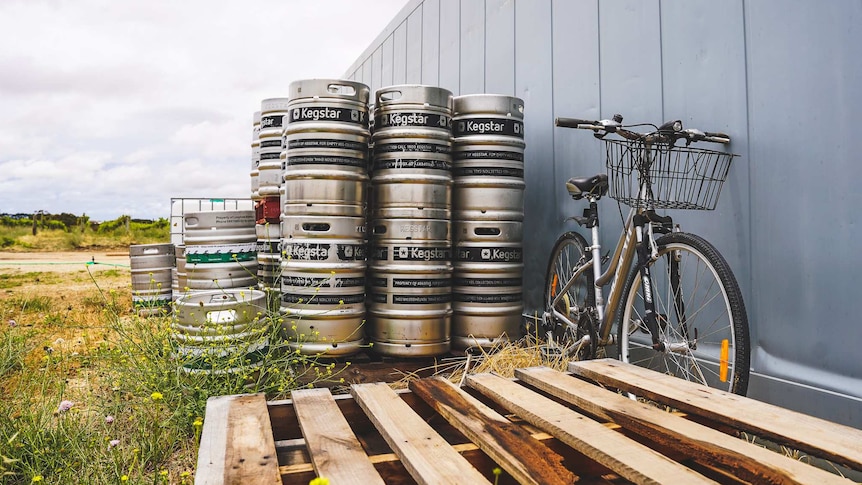 A bicycle rests outside against a large shed wall, several stacks of silver kegs behind it.