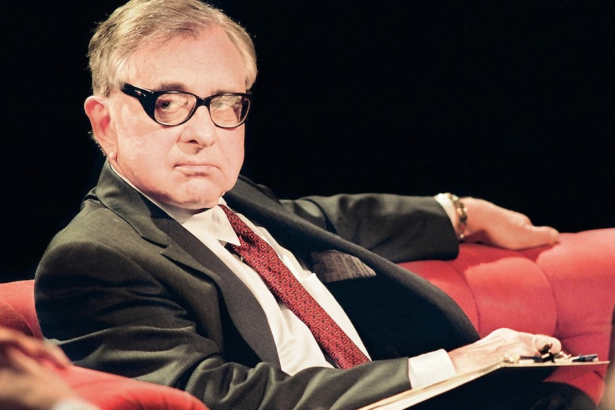 A man in glasses and a suit sits on a red couch