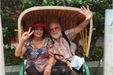 A woman making a peace sign and a man waving sit in a carriage smiling.