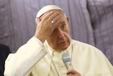 Pope Francis touches his forehead as he holds a microphone.