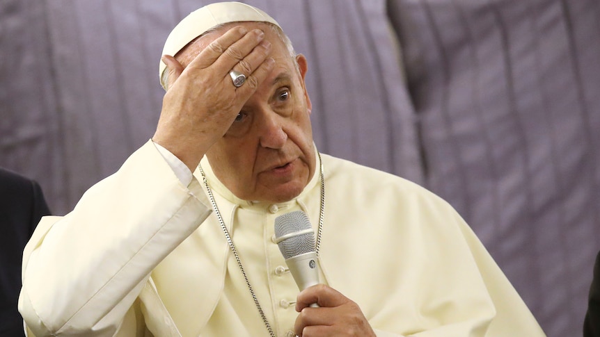 Pope Francis touches his forehead as he holds a microphone.