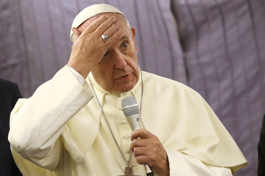 Pope Francis touches his forehead as he holds a microphone.