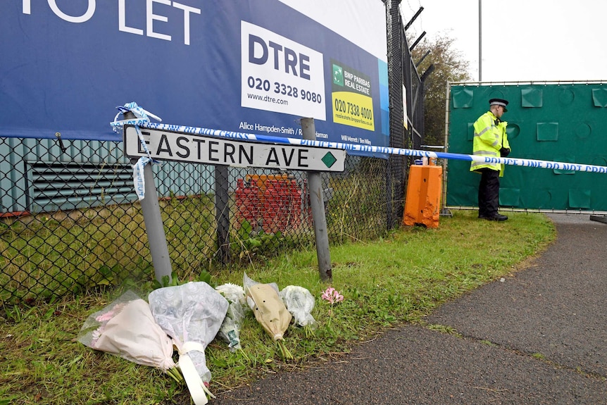 Five bouquets and one floral stem sit on grass in front of a sign reading "Eastern Avenue". A policeman is behind.