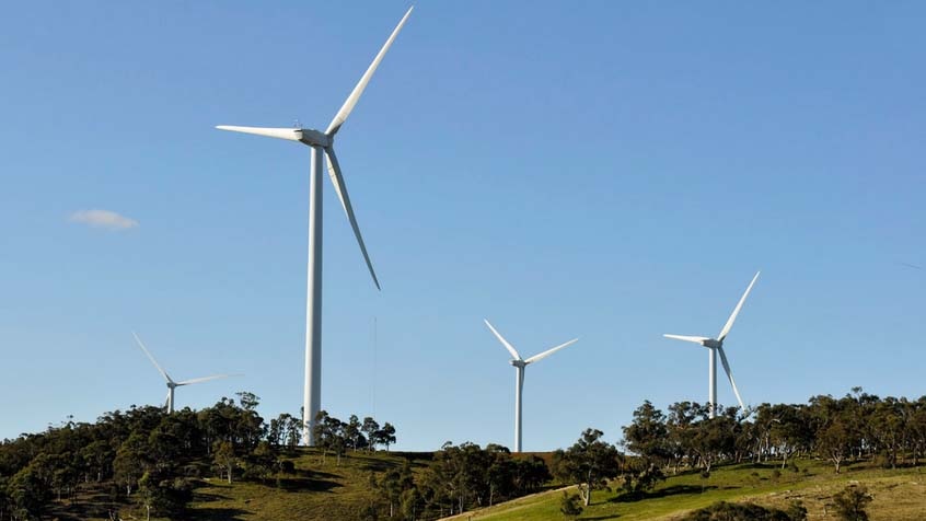 Four wind turbines jut out of a lush green hillside against a blue sky.