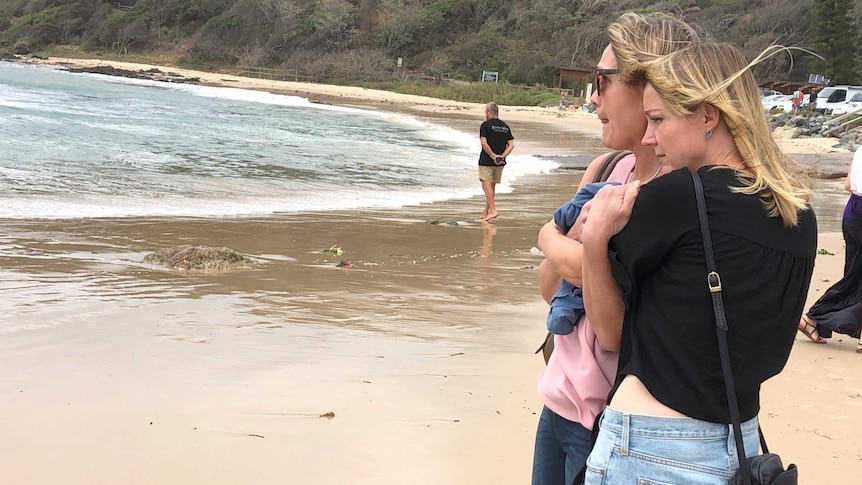 Two women, looking sad, stand at the edge of the ocean