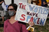 A woman wearing a face mask and sunglasses holds up a sign that says the voters have spoken