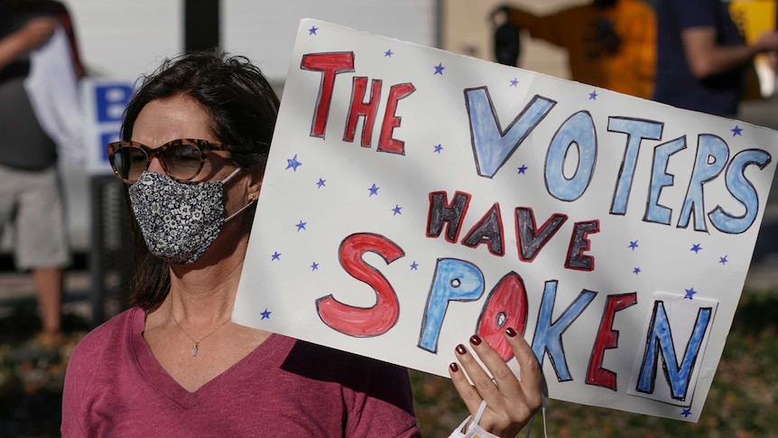A woman wearing a face mask and sunglasses holds up a sign that says the voters have spoken