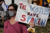 A woman wearing a face mask and sunglasses holds up a sign that says the voters have spoken