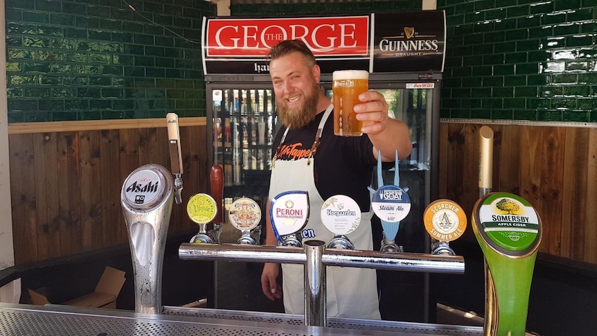 A man with a beard holds a beer behind a bar