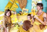 Three children, a woman and a dog wearing yellow sit in a loungeroom surrounded by objects that are also yellow.