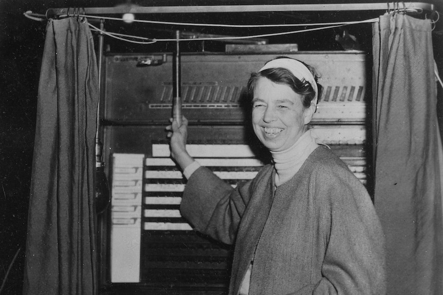 Elanor Roosevelt smiles as she holds the lever of a voting machine.