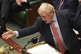 British Prime Minister Boris Johnson wears a blue suit and red tie and speaks into a microphone while gesturing across a table.