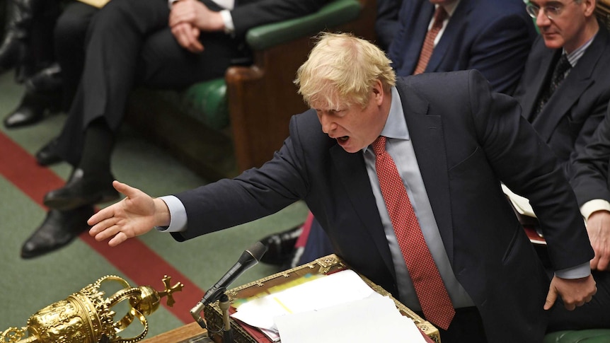 British Prime Minister Boris Johnson wears a blue suit and red tie and speaks into a microphone while gesturing across a table.