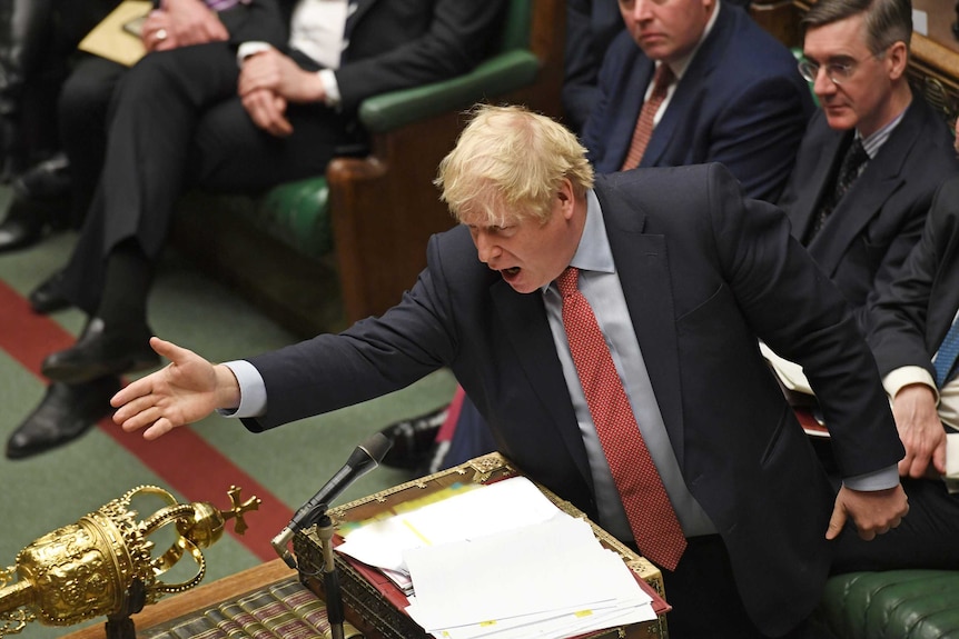 British Prime Minister Boris Johnson wears a blue suit and red tie and speaks into a microphone while gesturing across a table.