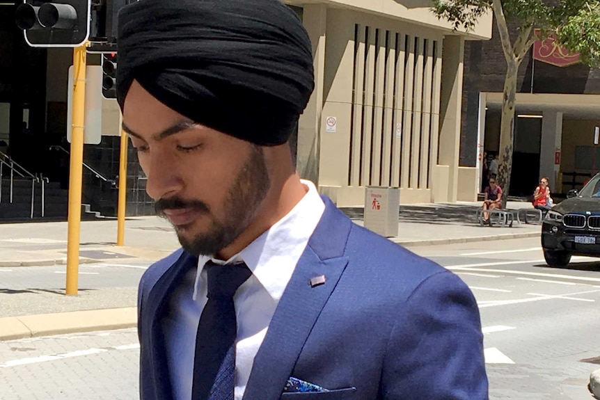 A young man wearing a blue suit and black turban walks on the street outside a court building.