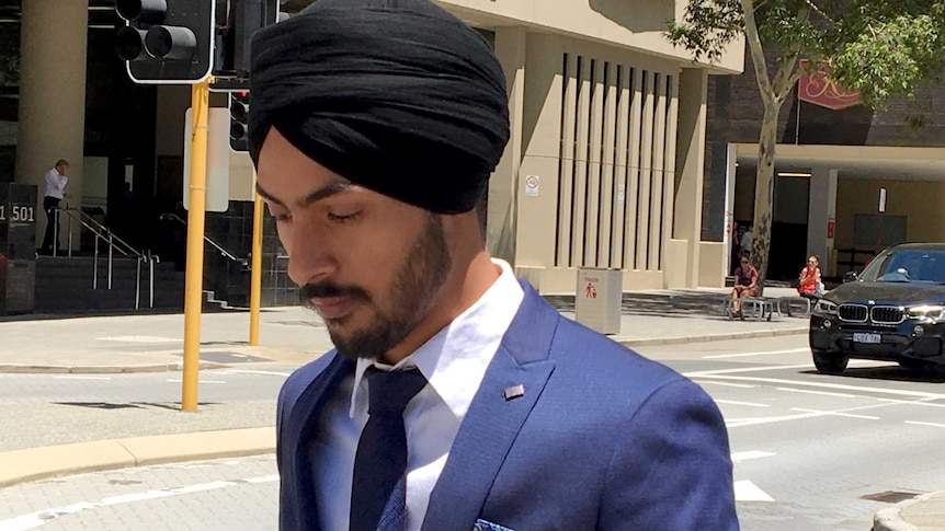 A young man wearing a blue suit and black turban walks on the street outside a court building.