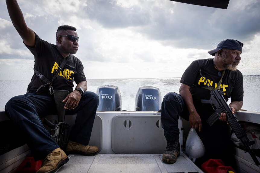 Two men wearing Tshirts with POLICE in yellow writing sit on the back of a boat out on the water