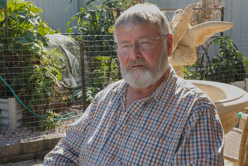 A greying man with glasses sits at outside garden table looking concerned