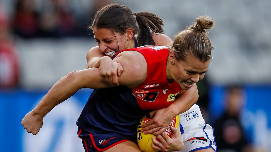 A Melbourne Demons AFLW player holds the ball as she is tackled by a North Melbourne opponent.