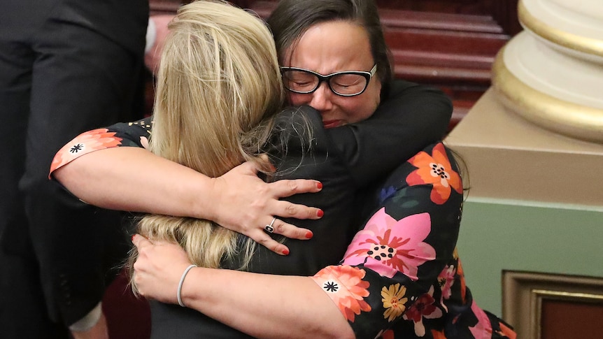 Two women firmly embrace one another, as one has her eyes closed as if she's crying.