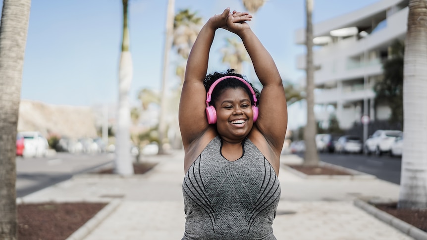 A woman wearing pink headphones and exercise gear stretches her arms above her head smiling