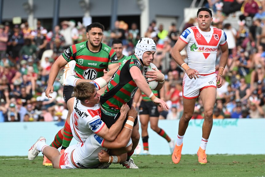 A South Sydney NRL player wearing headgear falls forward carrying the ball as he is tackled by several defenders.