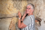 A woman with short hair and glasses looks at the camera smiling while removing some old wallpaper