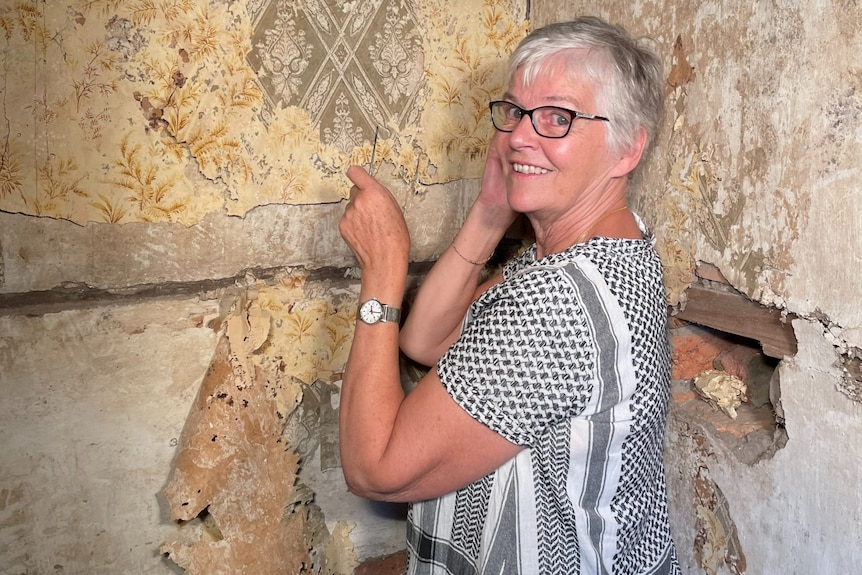 A woman with short hair and glasses looks at the camera smiling while removing some old wallpaper