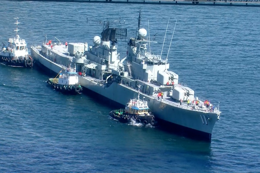 A large grey navy ship on water surrounded by three small tugboats