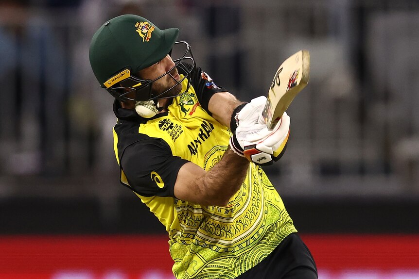A man in a green and gold Australian T20 cricket uniform and batting helmet is struck in the neck by a white cricket ball.