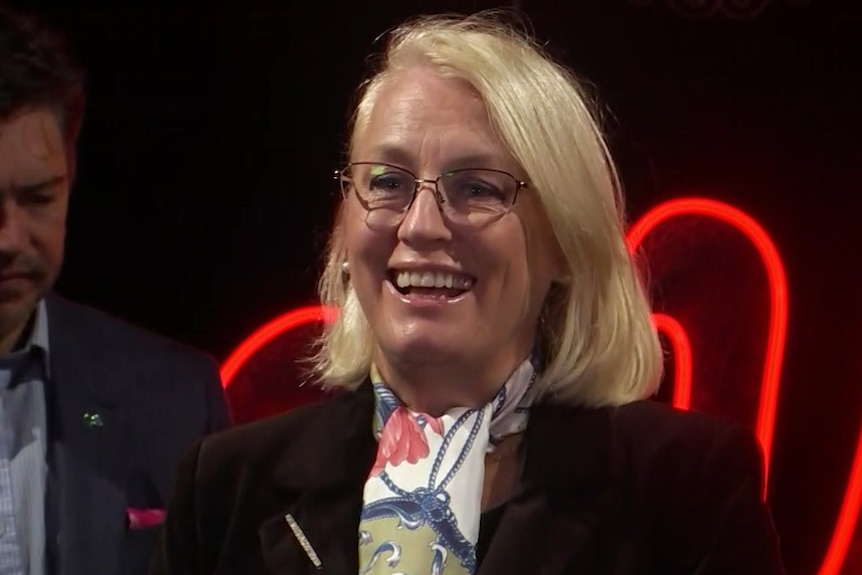 A smiling woman with blond hair and glasses speaking at a media conference.  