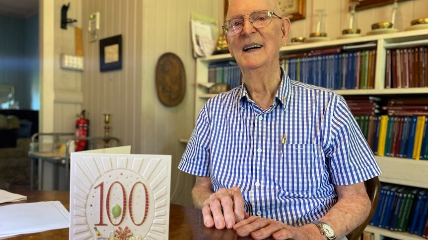 Dr John O'Hagan sits smiling at his dining table. A 100th birthday card is standing on the table in front of him.