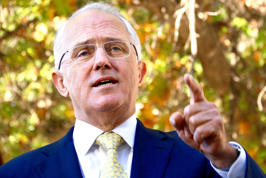 Malcolm Turnbull points as he speaks, with out of focus autumn leaves on trees in the background.
