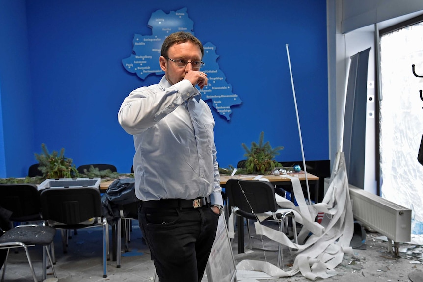 A man wearing a blue button-up shirt and black trousers puts his hand to his mouth while his office has debris from explosion.