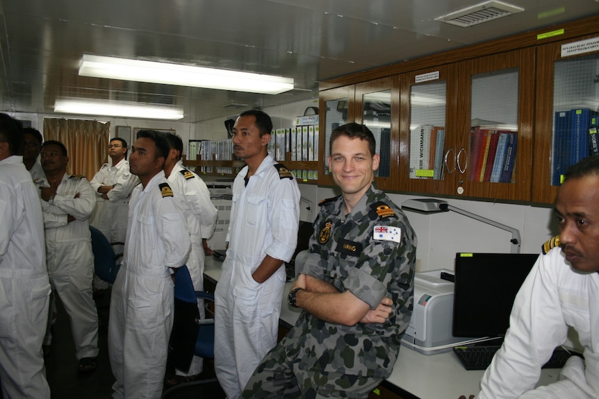 Australian Naval officer sitting back on a table with his arms folded. He's surrounded by Asian naval officers. 