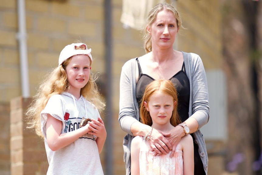 A mother stands with her arms around her daughter. A slightly older girl stands beside her holding a guinea pig.