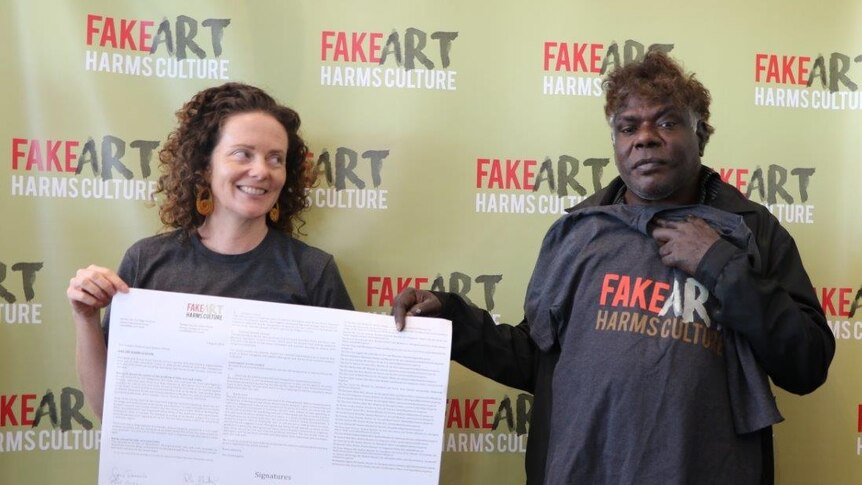 Gabrielle Sullivan and Gabriel Nodea hold up a petition in front of a Fake Art Harms Culture sign.