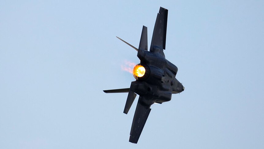 A dark grey fighter jet in a blue sky, on an angle, with a fiery blast of igniting jet fuel coming out the back.