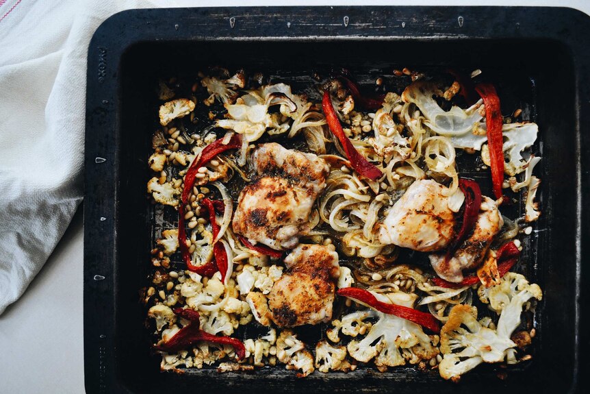 A baking tray with high sides with baked cauliflower, pine nuts, onion, chicken thighs and jarred capsicum, family dinner.