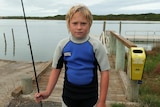 A young boy in a wetsuit holds a fishing rod but looks sad