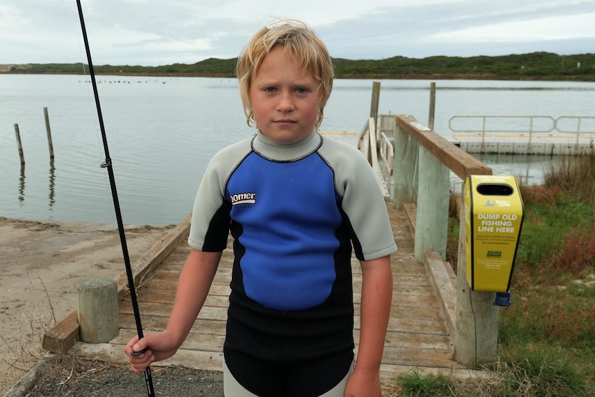 A young boy in a wetsuit holds a fishing rod but looks sad
