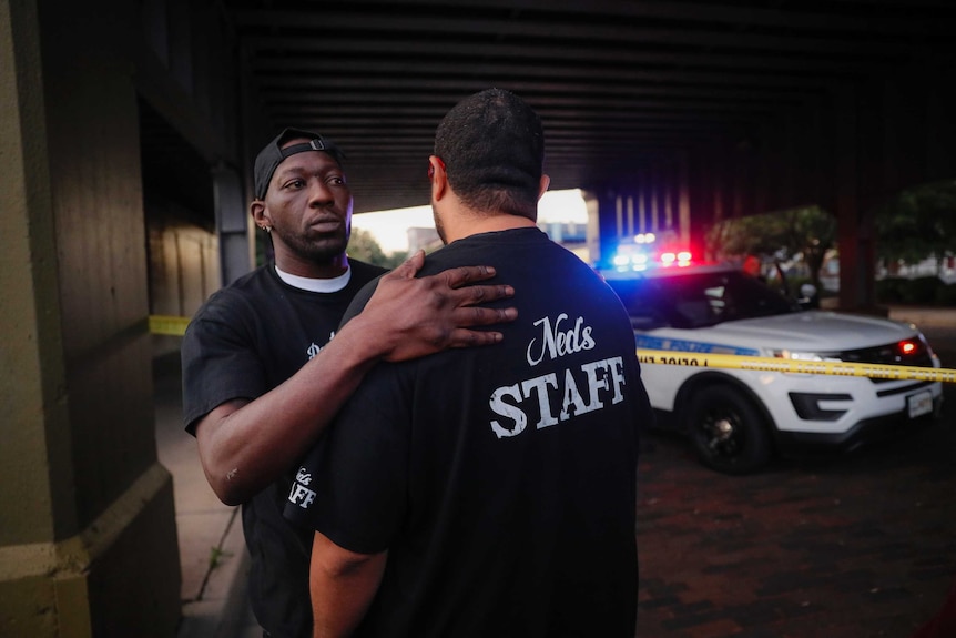 A man consoles another witness to a mass shooting in front of police tape and a patrol car