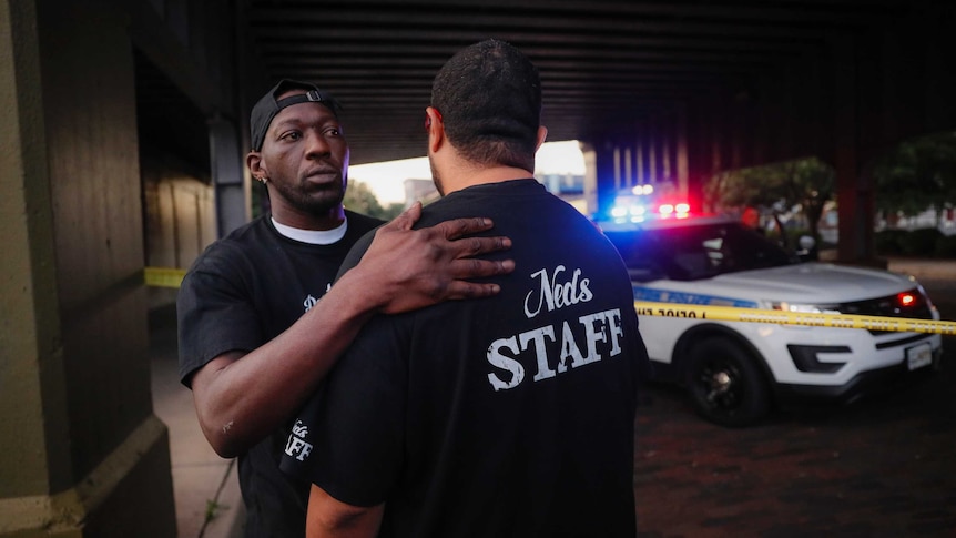 A man consoles another witness to a mass shooting in front of police tape and a patrol car