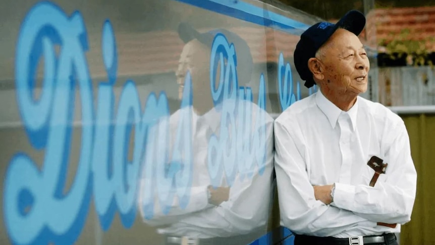 An elderly Asian main wearing a cap, stands with arms crossed and leans against a bus, while laughing. 