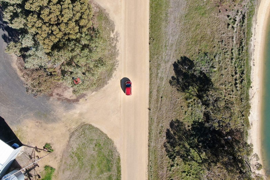 A drone shot shows a small red car travelling along a dirt road surrounded by trees and a dam in country Australia.