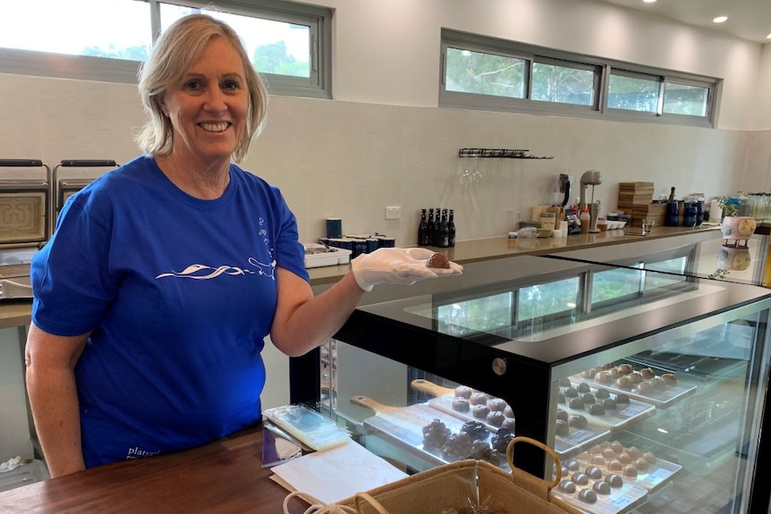 A woman with blonde hair and a blue t-shirt stands behind a glass display cabinet of chocolates holding a small chocolate.